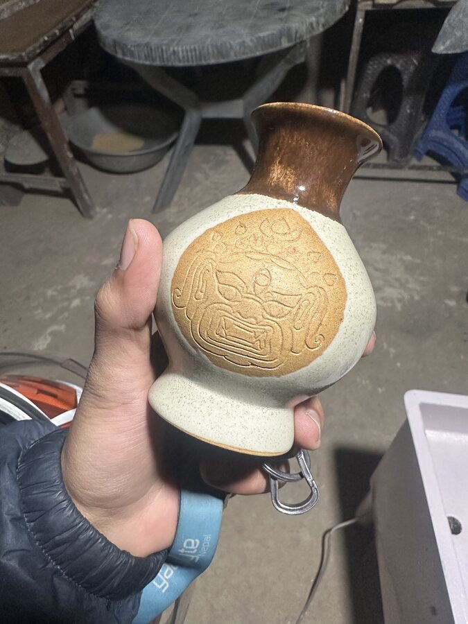 A hand holding a glazed clay pot with a carved face